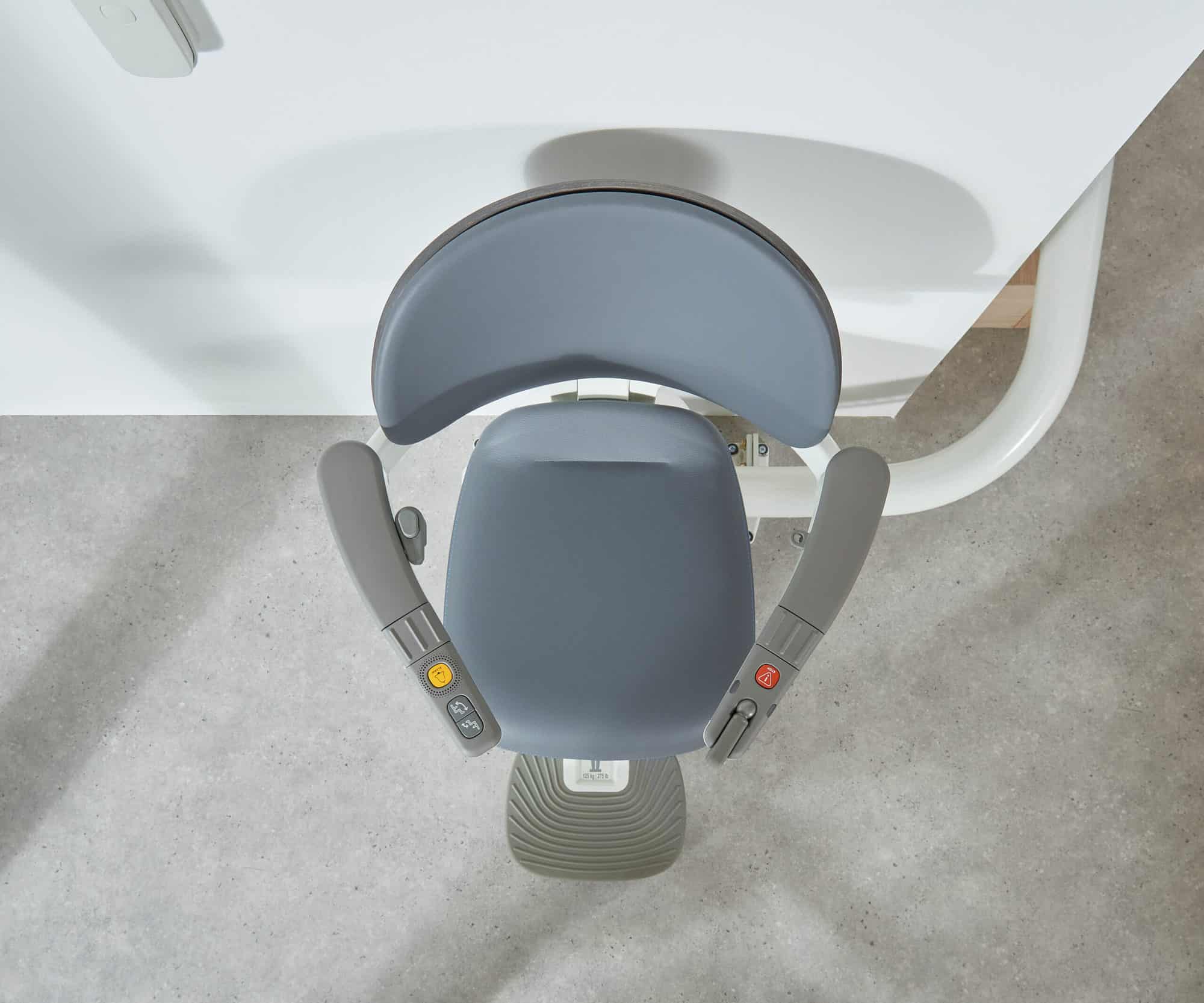 An installed Flow X stairlift with a teal seat positioned at the bottom of a staircase. The rail runs neatly along the side of the black stairs, with a small table and lamp beside it in a modern hallway.