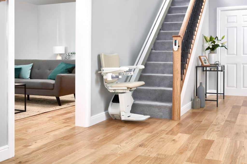 The Handicare 1100 stairlift positioned at the base of a staircase, shown with its slim rail and compact folded seat in a neutral-coloured hallway.